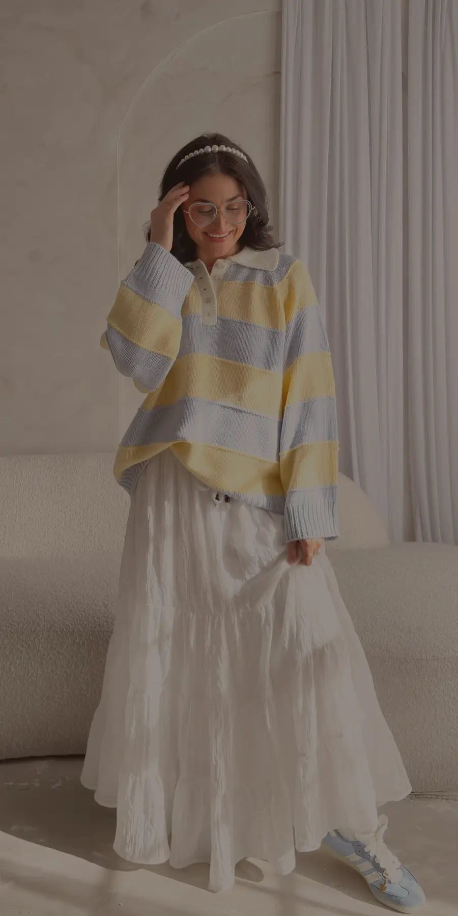 Woman styling a pastel yellow and blue striped knit sweater with a white collared neckline, paired with a feminine white tiered maxi skirt and light blue sneakers in a bright, airy room.