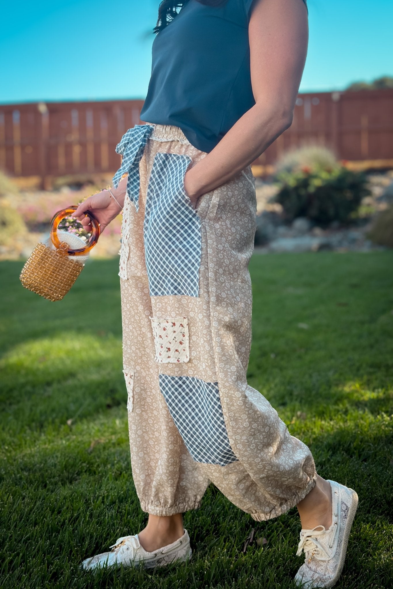Woman wearing beige floral patchwork balloon pants with navy gingham panels, navy top, straw hat and white sneakers for an easy transitional outfit.