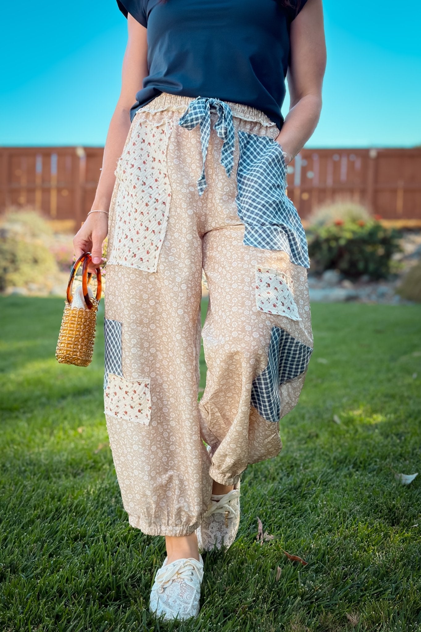 Woman wearing beige floral patchwork balloon pants with navy gingham panels, navy top, straw hat and white sneakers for an easy transitional outfit.