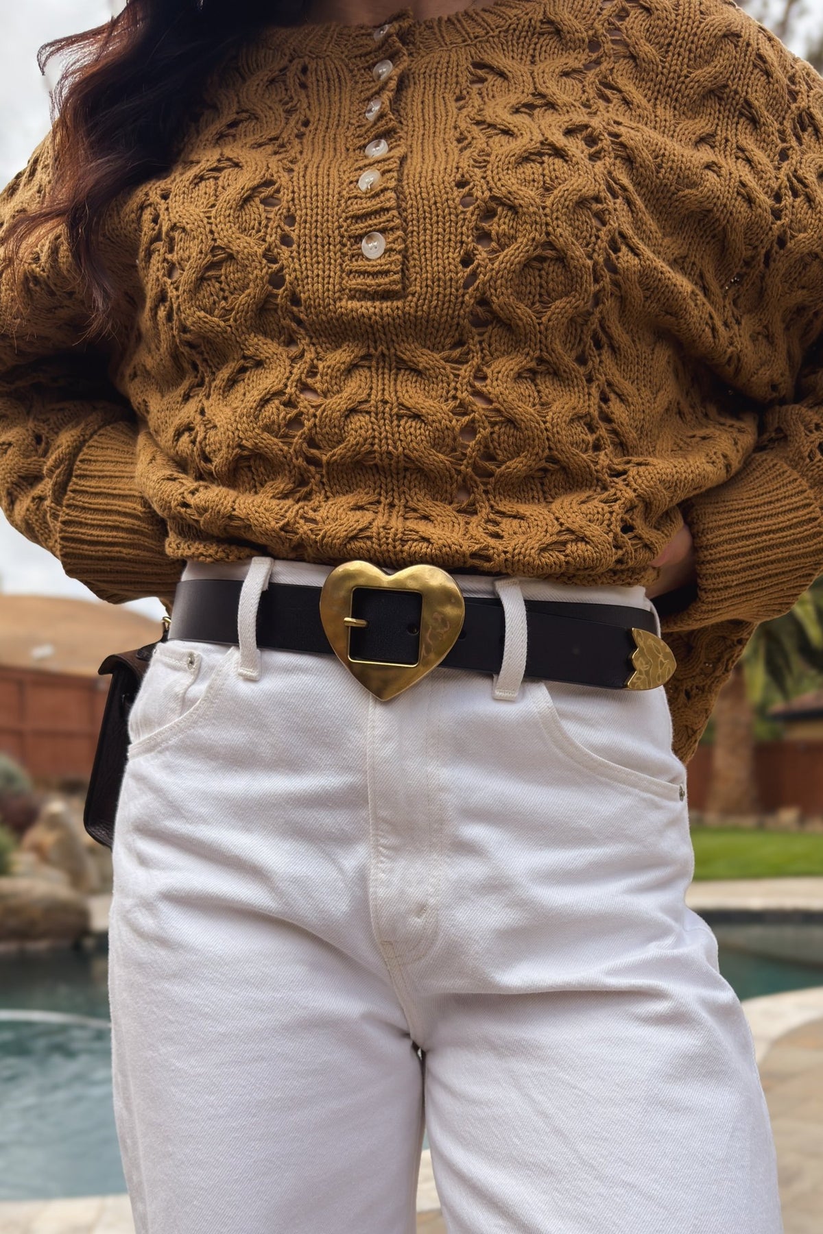 Black leather belt with oversized gold heart buckle worn with white denim and a warm brown open-knit sweater.