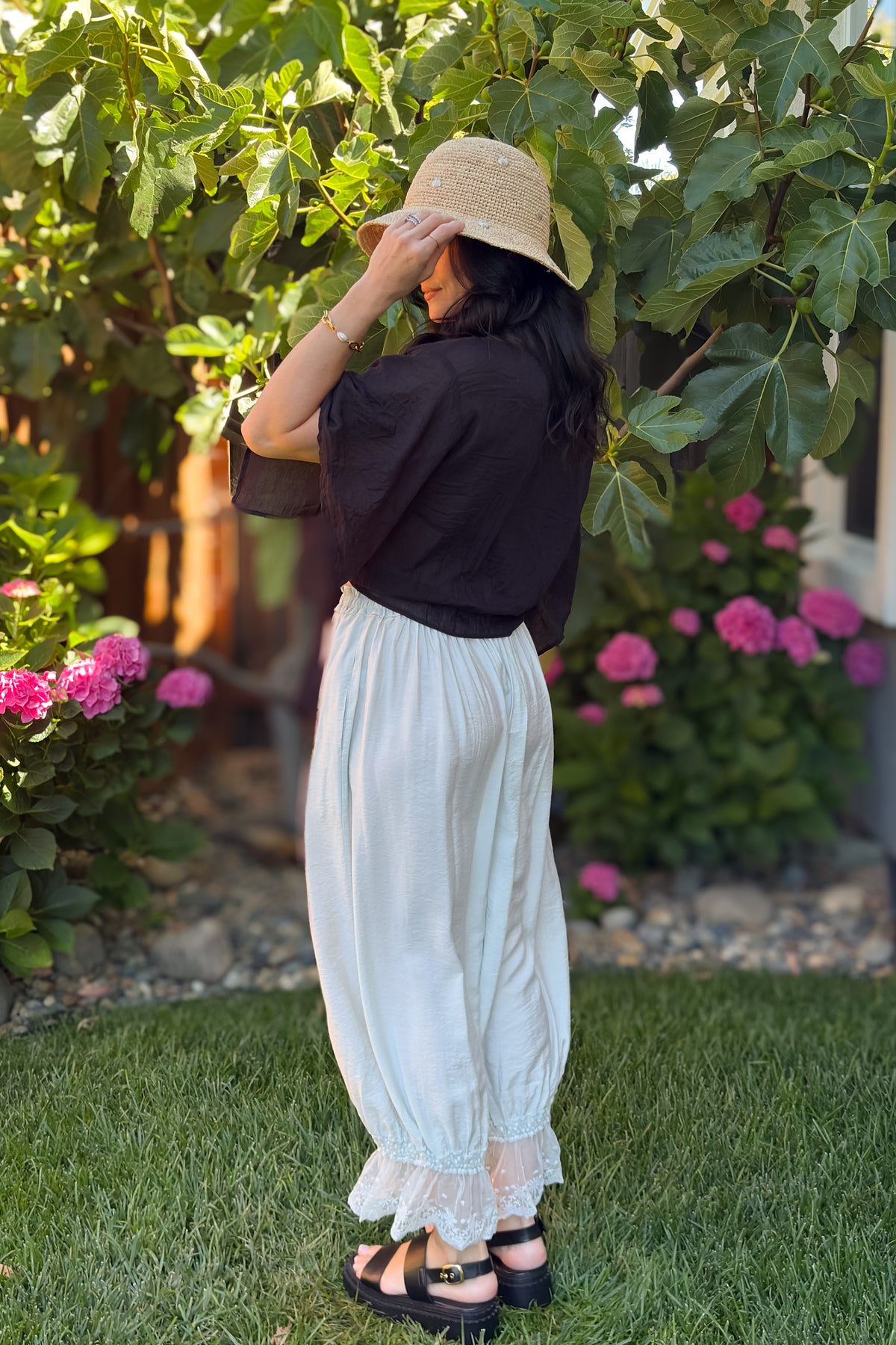 Back view of a black tie front kimono cover up top styled with cream lace hem pants and a straw hat.