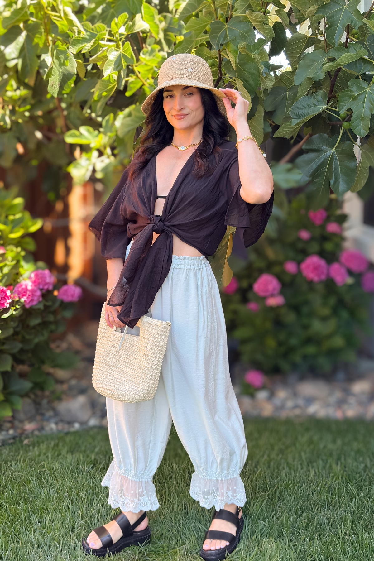 Black tie front kimono cover up top styled with cream lace hem pants, woven handbag, and straw hat in a feminine vacation outfit.