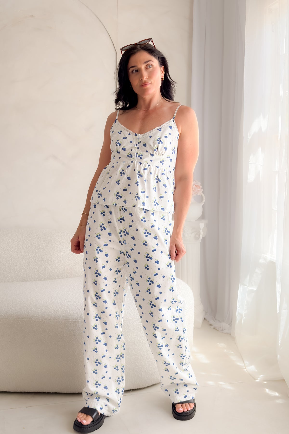 Woman wearing a white blueberry print babydoll tank top with lace trim neckline, adjustable straps, and feminine gathered silhouette.