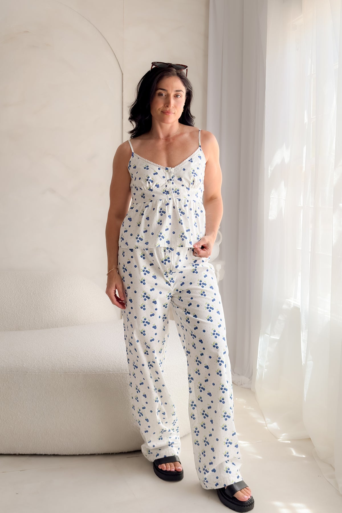 Woman wearing a white blueberry print babydoll tank top with lace trim neckline, adjustable straps, and feminine gathered silhouette.