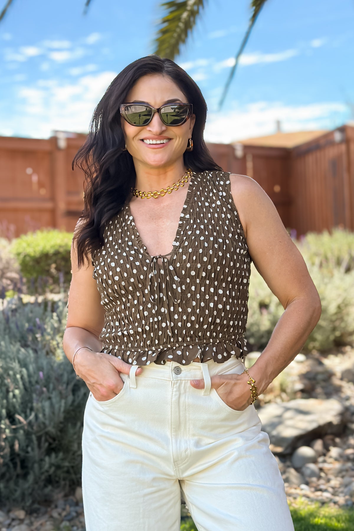 Side view of a sleeveless brown polka dot top with an adjustable front tie and fitted smocked waist.
