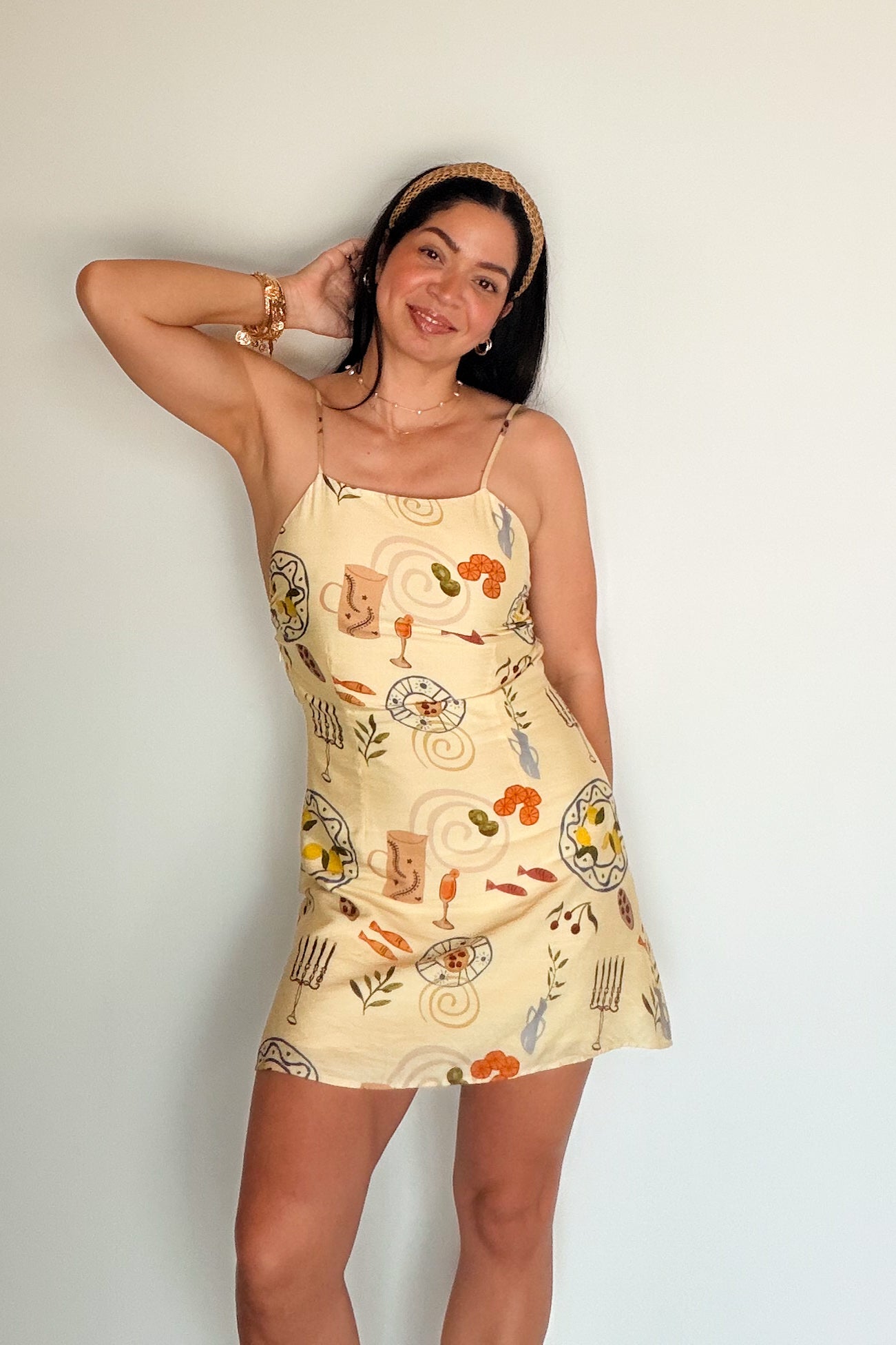 Woman wearing a butter yellow printed mini dress with spaghetti straps, styled with a woven headband and gold jewelry.