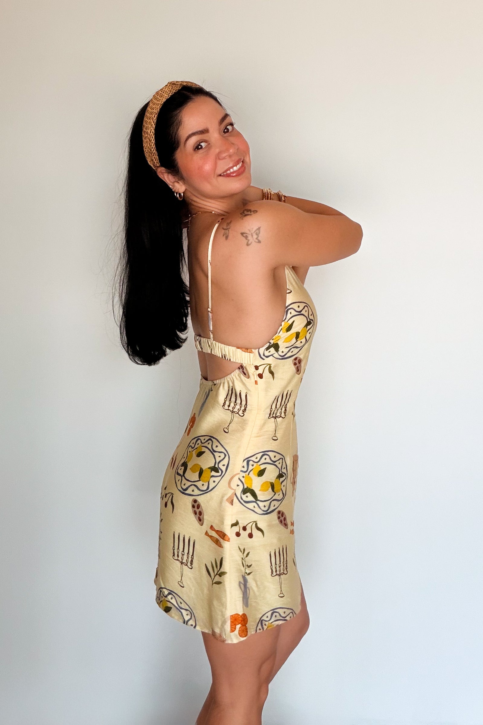 Woman wearing a butter yellow printed mini dress with spaghetti straps, styled with a woven headband and gold jewelry.