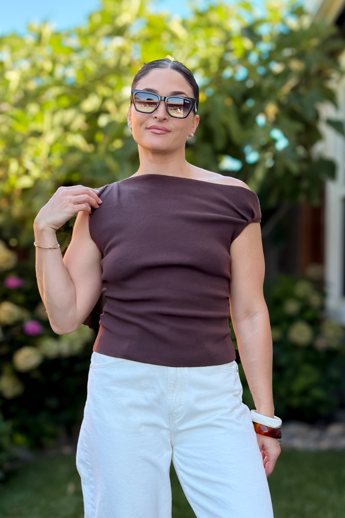 Woman wearing a chocolate brown off shoulder knit top with white wide leg jeans, showing a soft fitted silhouette and elegant everyday styling.