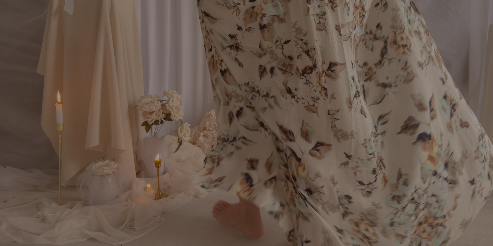 Close-up of a flowy floral summer dress in soft neutral tones with movement, styled in a romantic setting with candles, draped fabric, and delicate florals