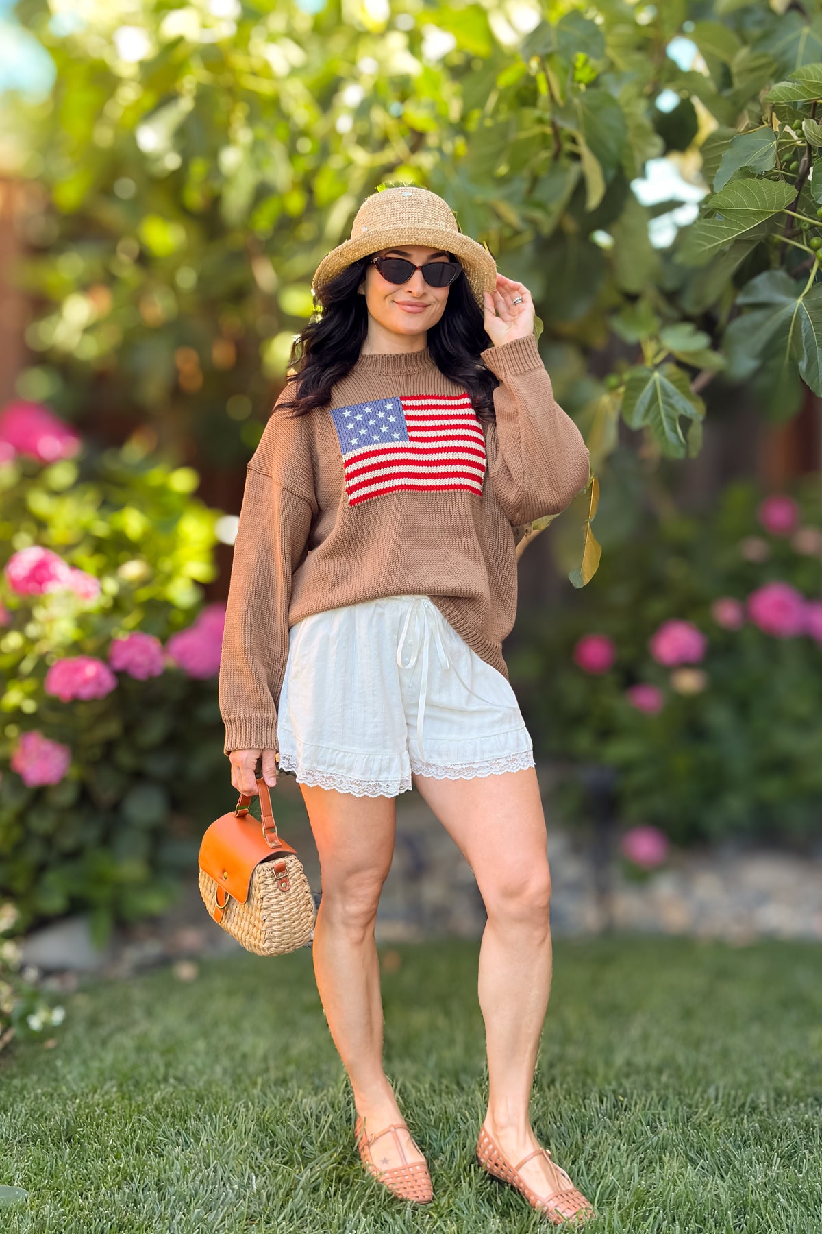 Women's brown American flag sweater styled with ivory lace shorts, straw hat, and bangles.