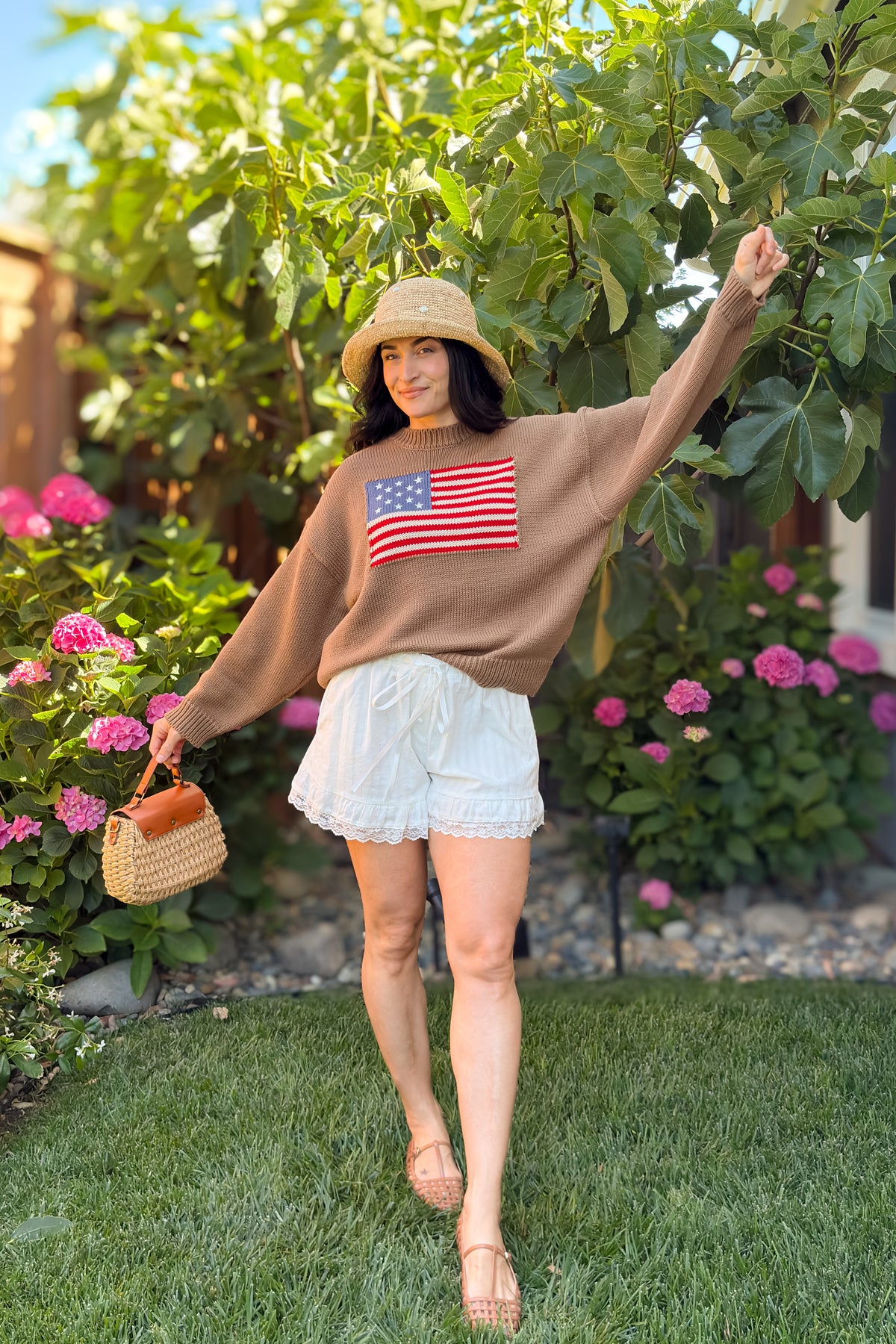 Women's brown American flag sweater with ivory lace trim shorts, straw hat, and woven handbag.