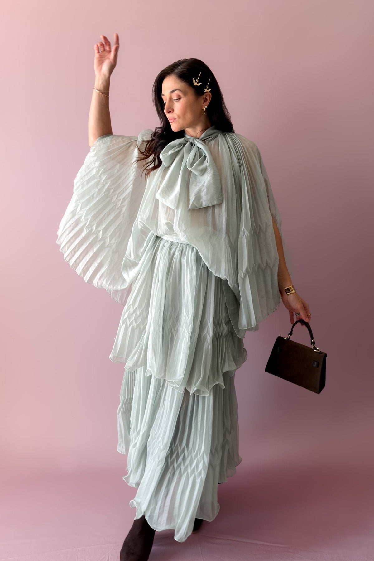 Woman wearing a mint pleated organza top and matching layered maxi skirt set with a dramatic bow neckline, styled with a structured brown handbag.