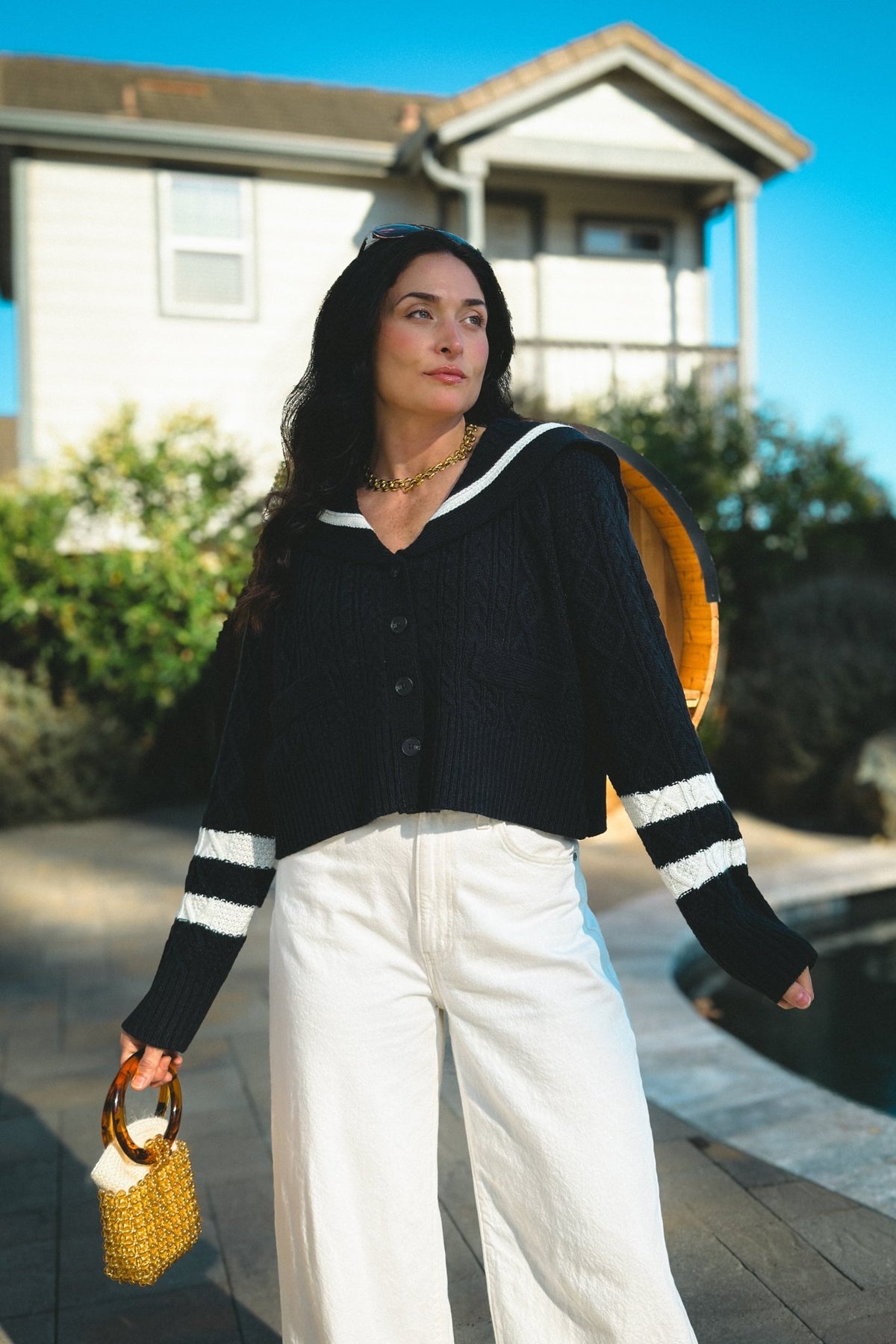 Close-up outfit view of a navy cable knit cardigan sweater with contrast white sailor collar and striped cuffs, paired with white denim and a gold beaded handbag.