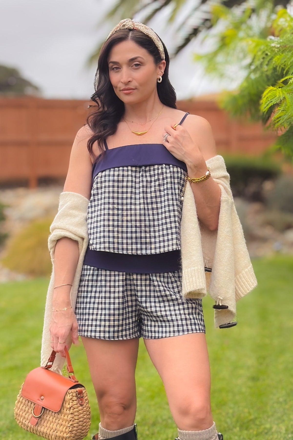 Woman wearing a navy and ivory plaid cami and shorts set with foldover details, styled with tall black boots, knit socks and a cream cardigan.