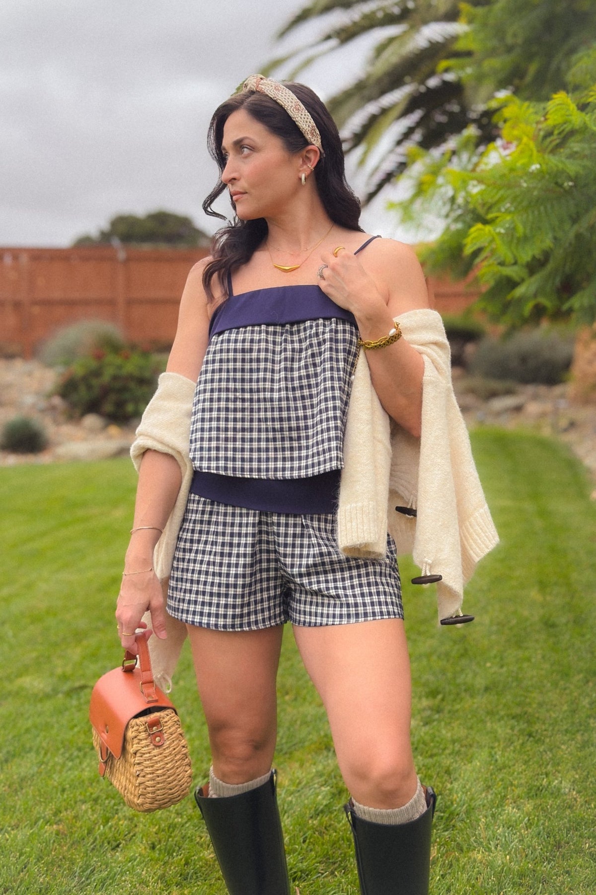 Woman styling a soft navy plaid cami short set with a cream knit cardigan draped over the shoulders and tall black boots.
