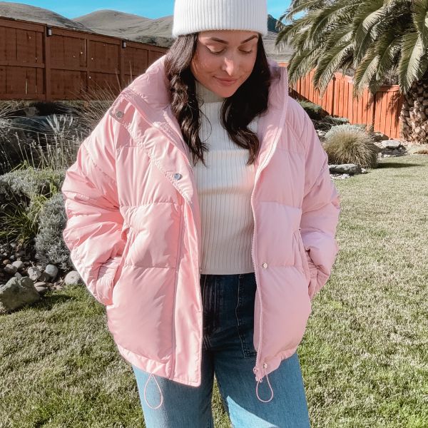 A brunette wearing a pink puffer coat while smiling and standing outside on green grass