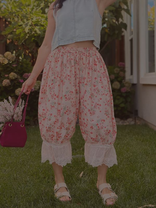 Woman wearing pink floral lace-trim bloomer pants with a soft drawstring waist, holding a burgundy handbag in a garden setting.