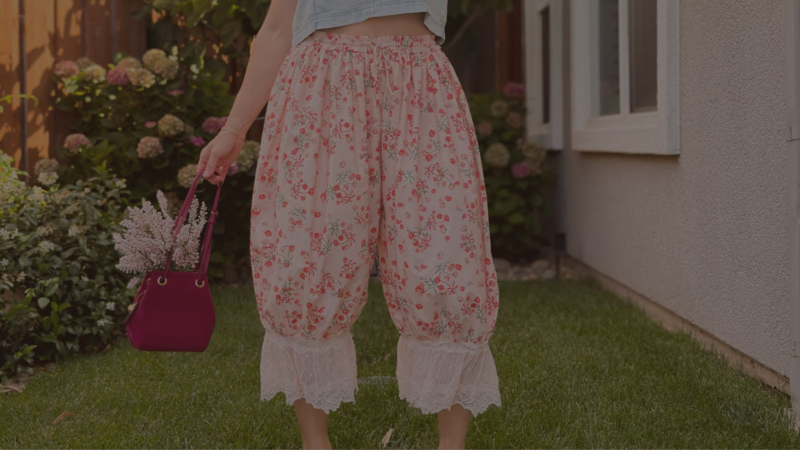 Woman wearing pink floral lace-trim bloomer pants with a soft drawstring waist, holding a burgundy handbag in a garden setting.