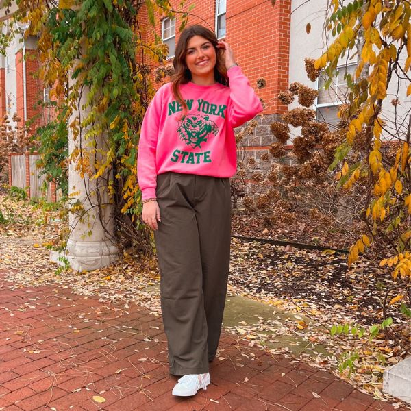 A brunette woman wearing a hot pink Daydreamer Graphic Sweatshirt and Olive Trousers with her hand in her hair