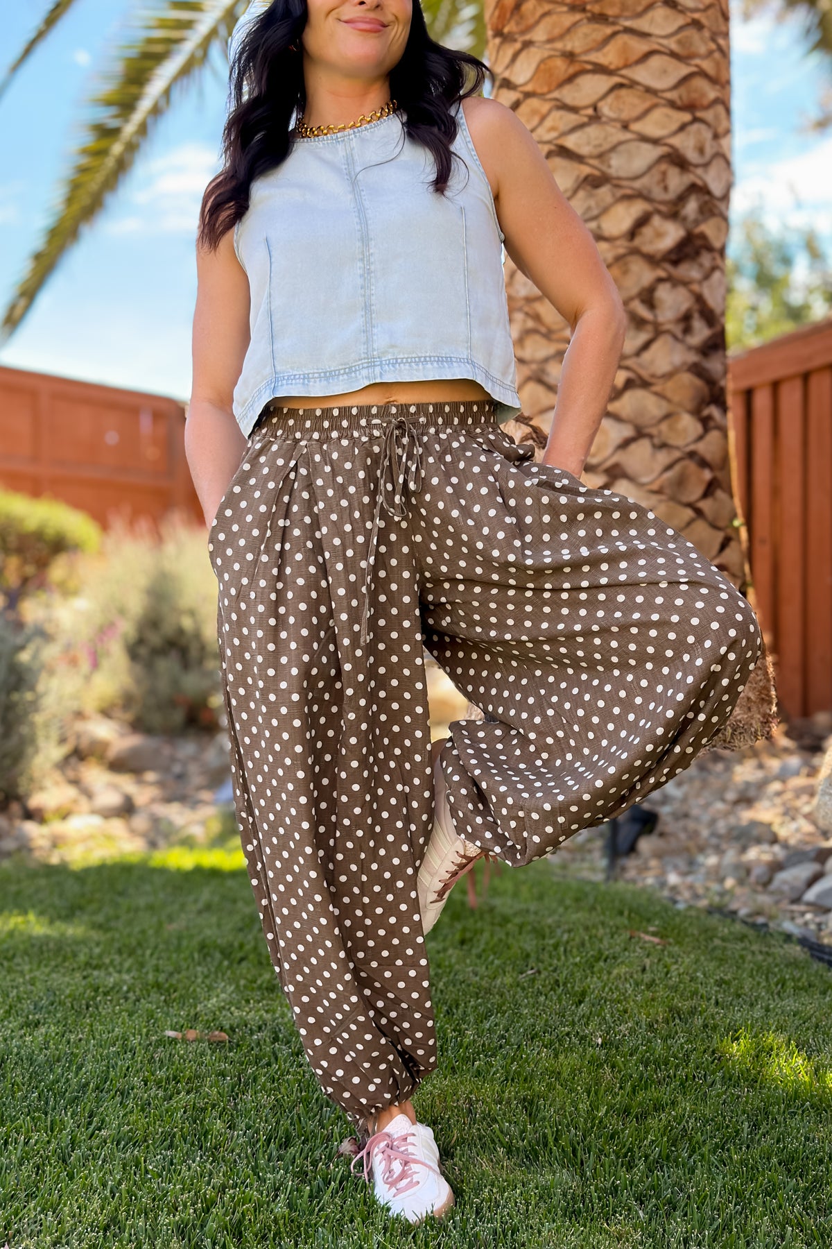 Woman wearing washed brown polka dot balloon pants with a light blue cropped denim tank and sneakers.